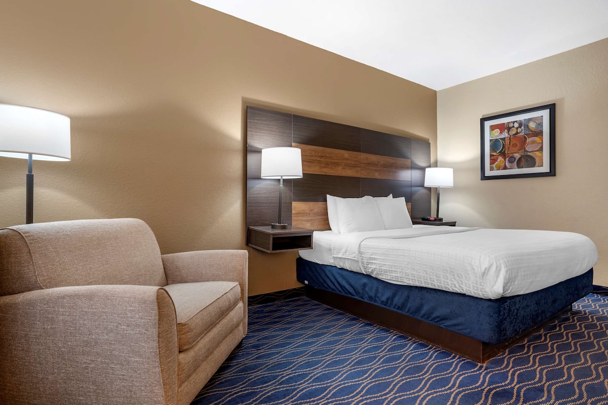 A hotel room with a bed, two bedside lamps, a chair, and a framed artwork on the wall.