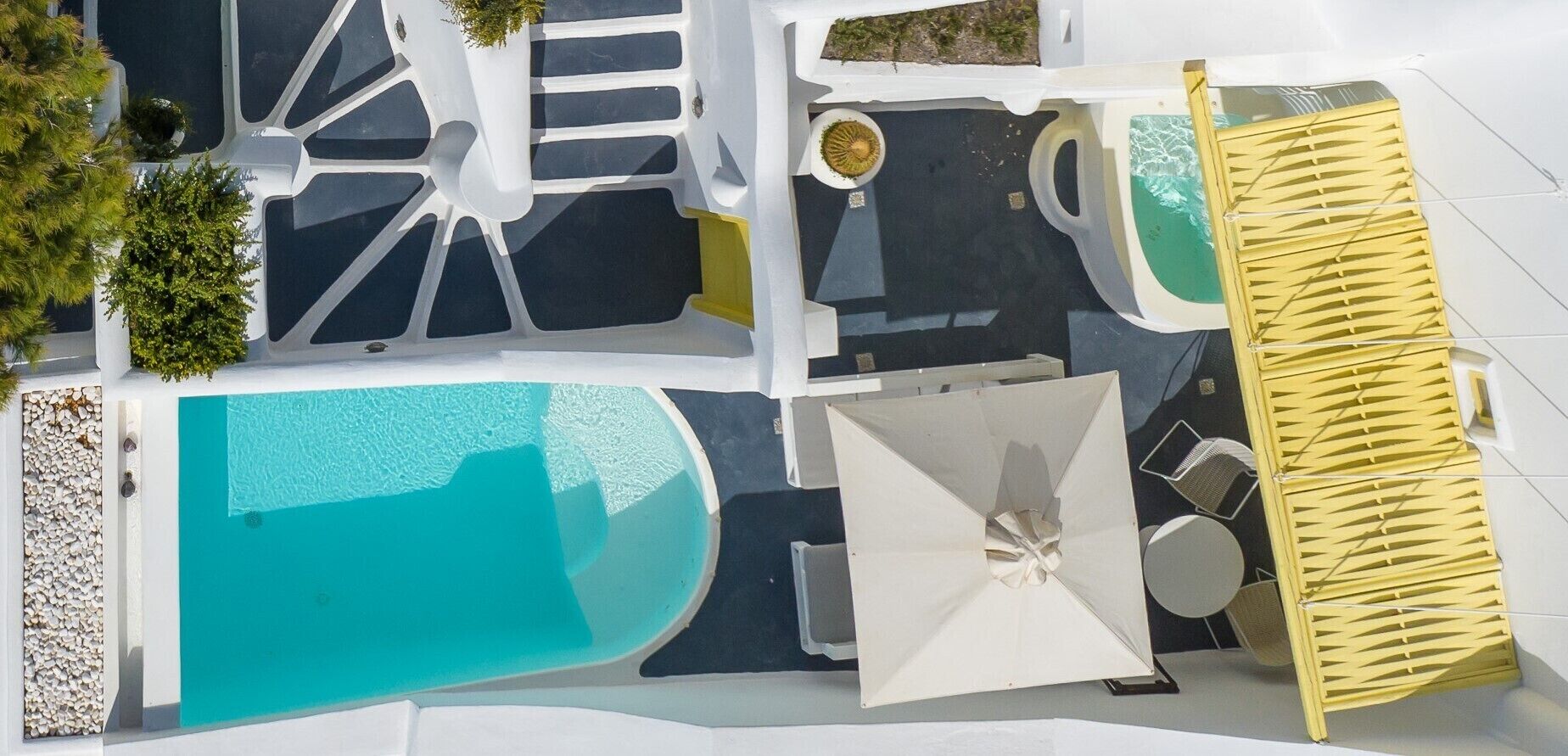 An aerial view of a modern pool area with multiple swimming pools, a sun lounger, and a small garden area.