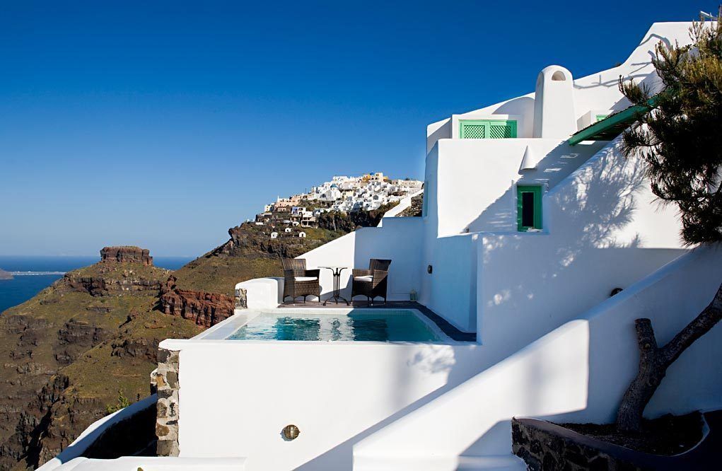 Un bâtiment blanc doté d’une piscine et d’un espace extérieur aménagé, offrant une vue sur un paysage côtier caractérisé par des bâtiments blancs et des falaises.