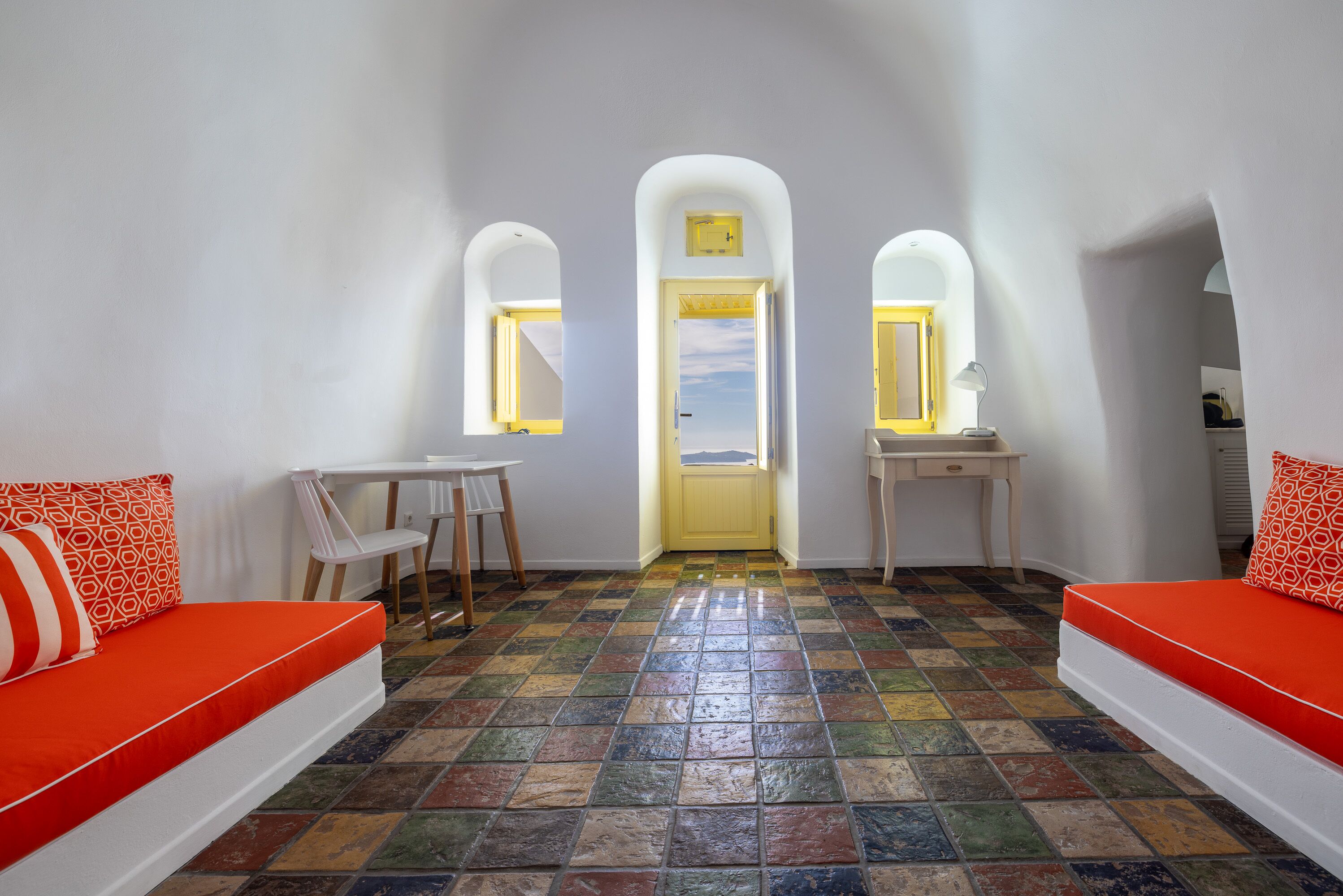 A room with a colorful tiled floor, a white sofa with orange cushions, a small table, and a chair.