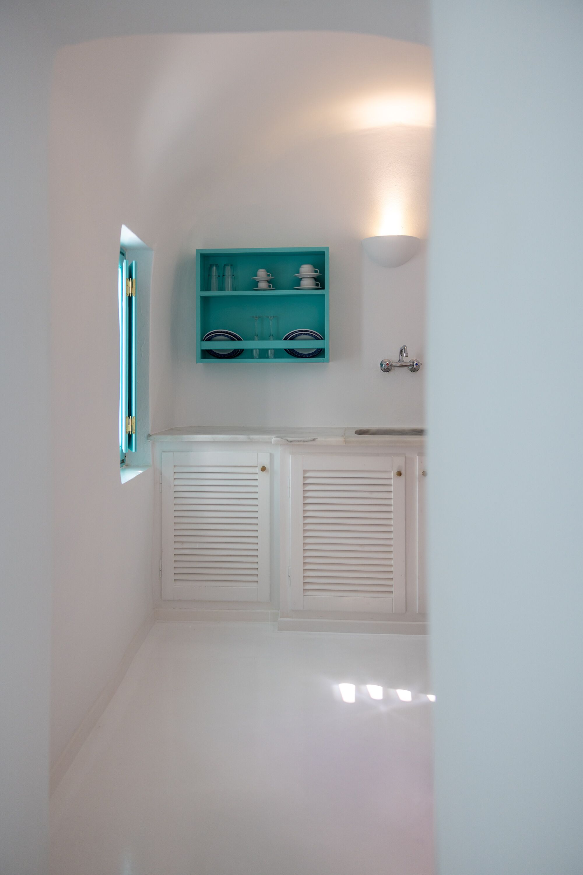Une salle de bain aux murs blancs, une fenêtre avec des stores, un radiateur blanc et une étagère turquoise où sont rangés les articles de toilette.