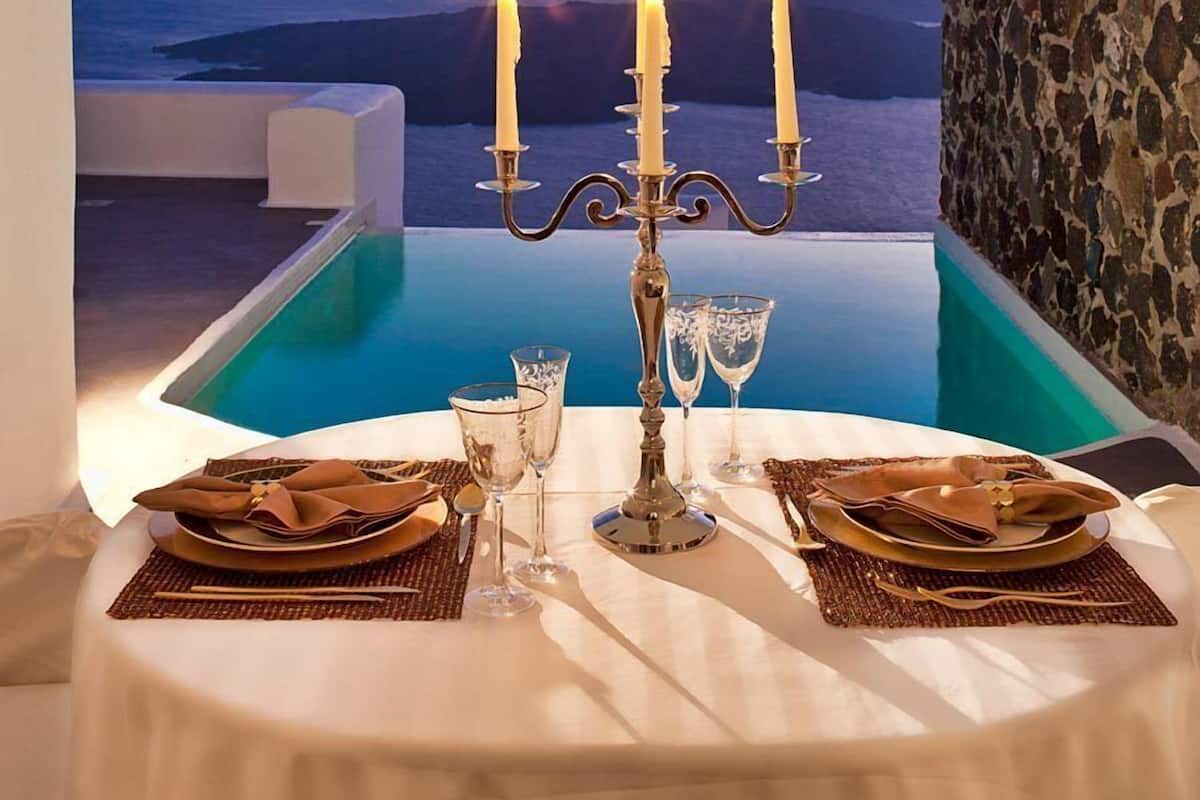 A table set for two with a candle centerpiece, overlooking a pool and a scenic view of the sea and hills.