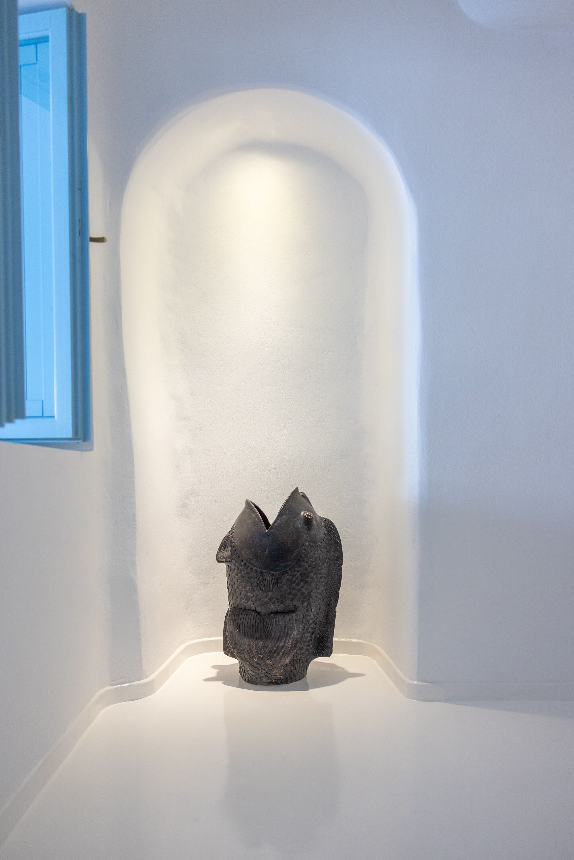 Une sculpture minimaliste, placée dans un coin d’une pièce aux murs blancs et dont le cadre de fenêtre est bleu.