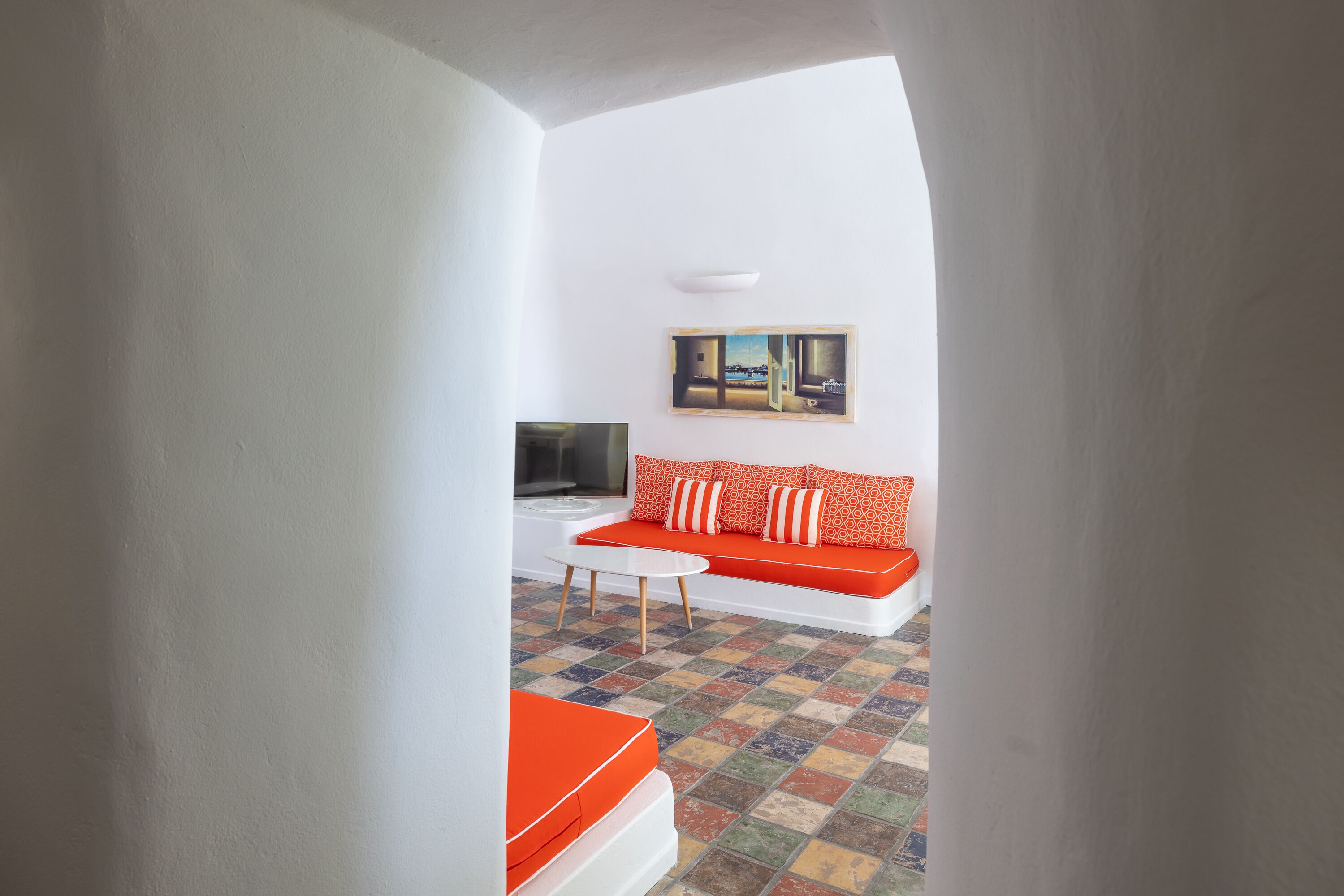 Un salon moderne avec un canapé blanc, des coussins orange, une petite table blanche et un sol carrelé aux couleurs vives.