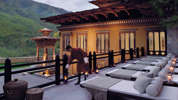 A traditional building with a tiled roof and wooden beams, a person tending to a lantern, and a row of lounge chairs with cushions and candles.