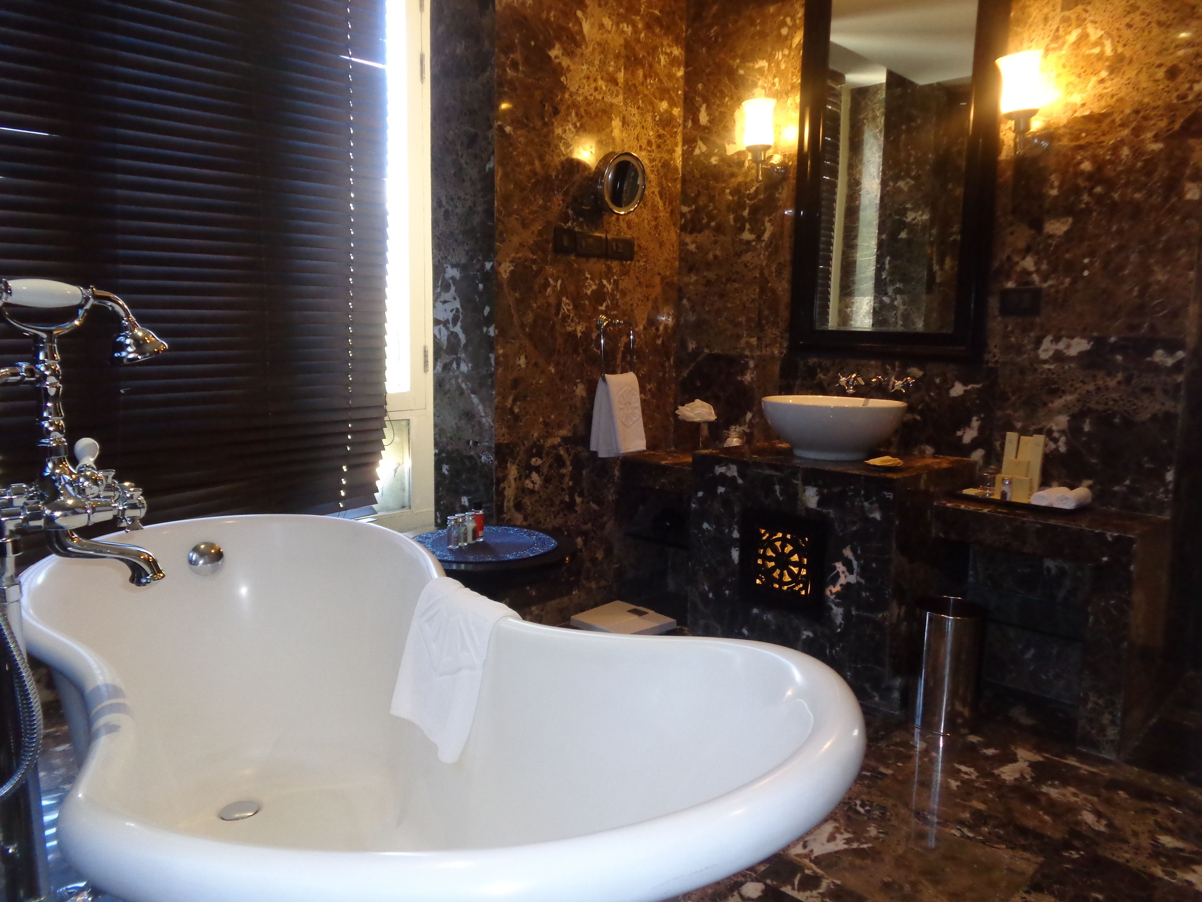 A bathroom with a large white freestanding bathtub, a round mirror, a white sink with a marble countertop, and a wall-mounted light fixture.