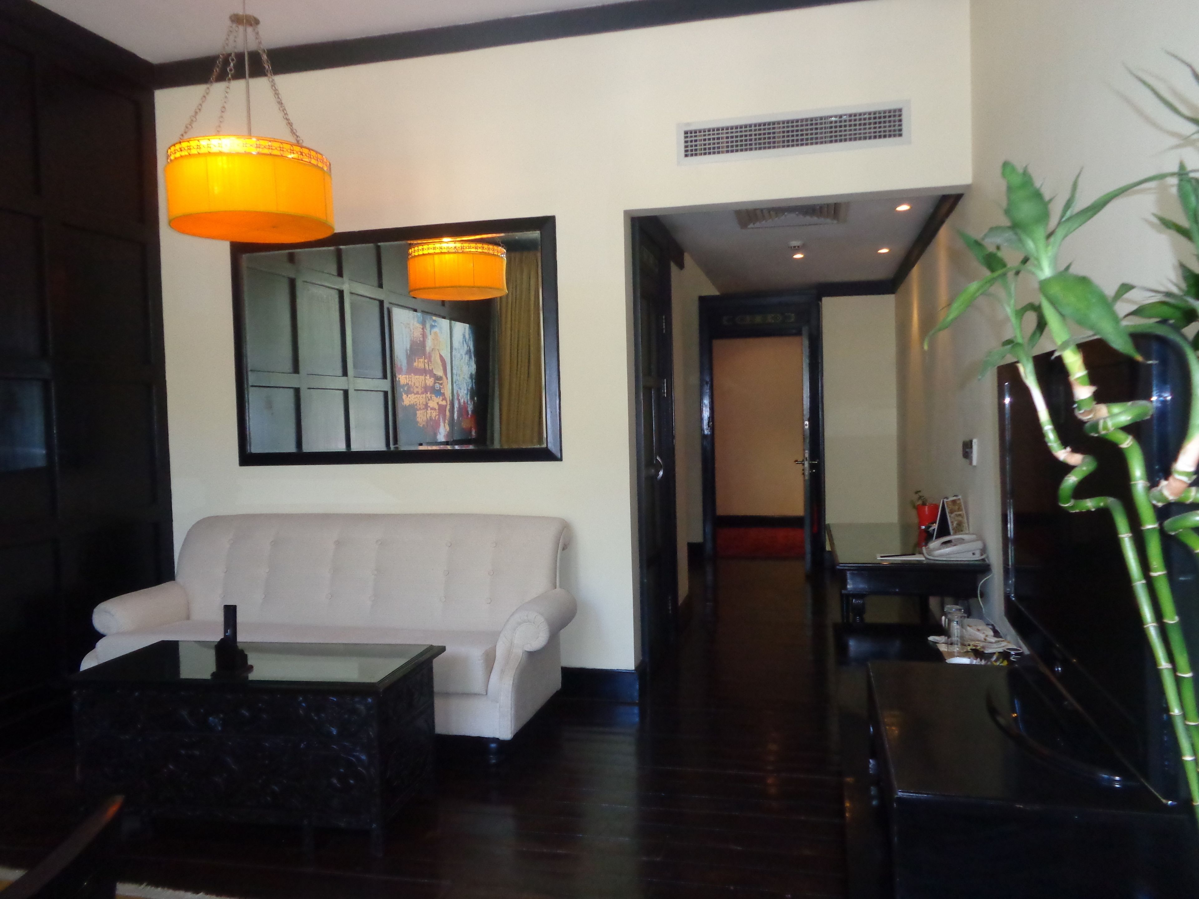 A modern living room with a white sofa, a glass coffee table, and a black side table. There is a large mirror on the wall and a hanging orange lamp.