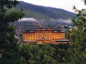A traditional building with multiple levels and a prominent central entrance, surrounded by lush greenery and mountains in the background. - Pemako Thimphu (Thimphu)
