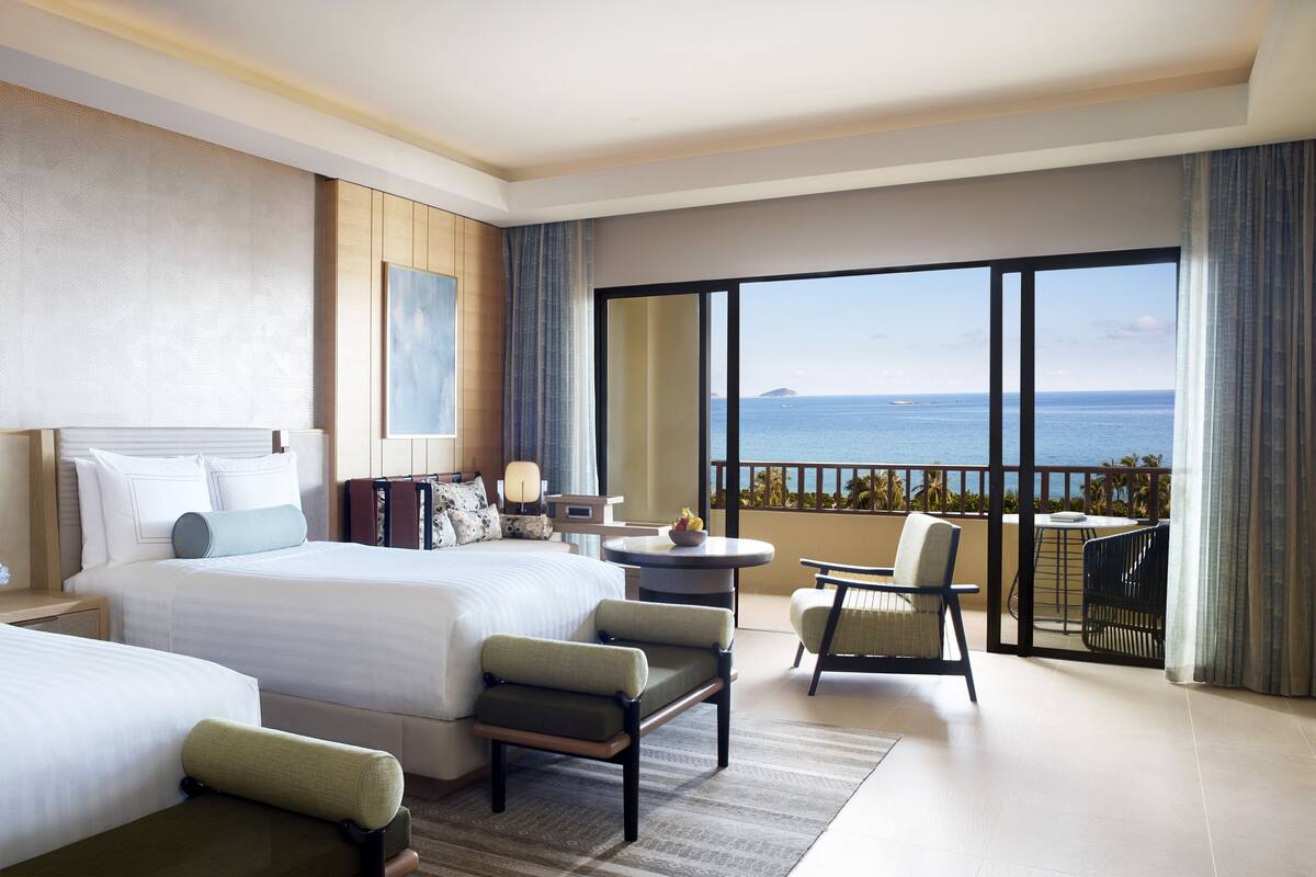 A hotel room with two beds, a balcony with a view of the sea, and a seating area with a table and chairs.