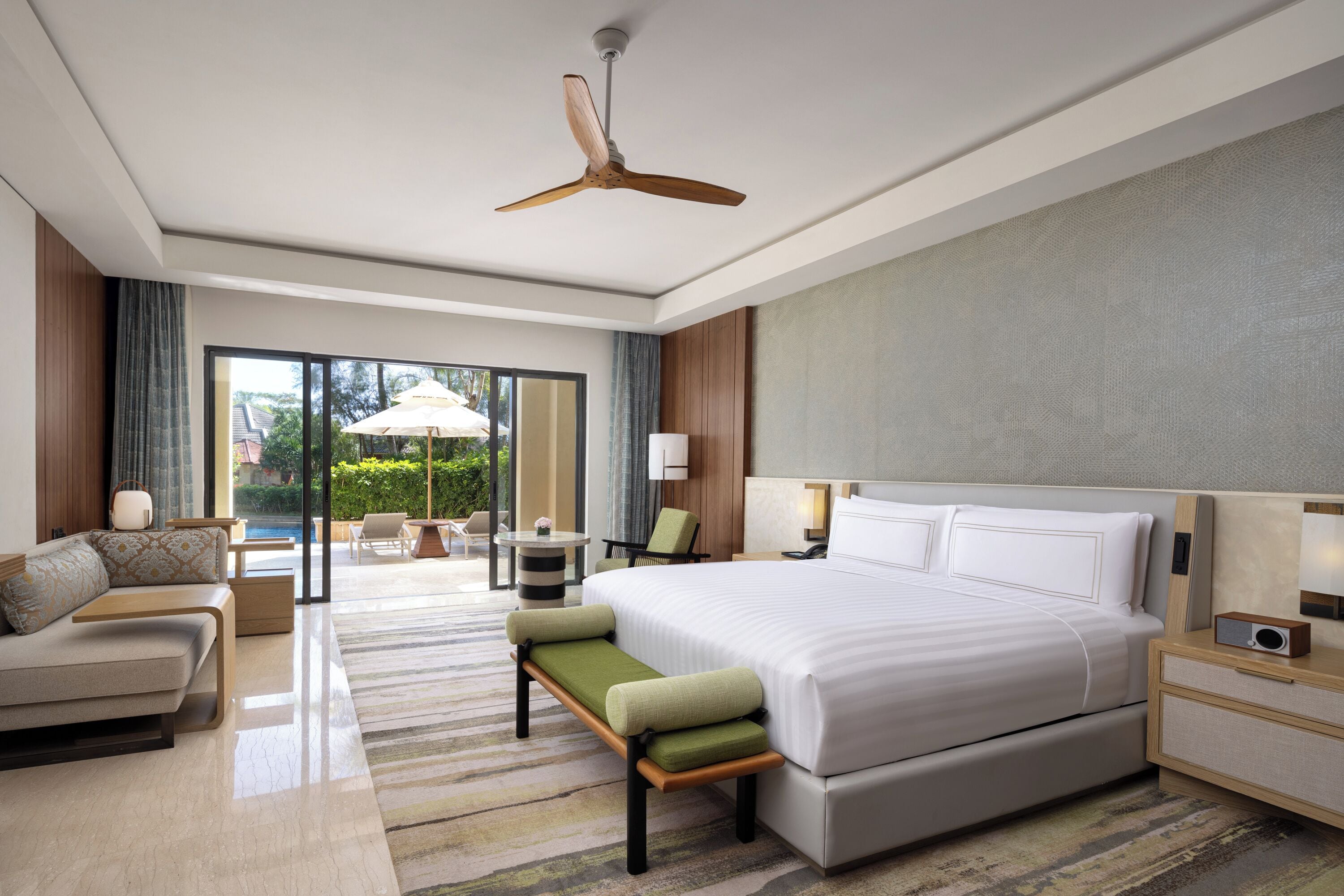 A modern hotel room with a large bed, a sofa, a bench, a desk, and a ceiling fan.