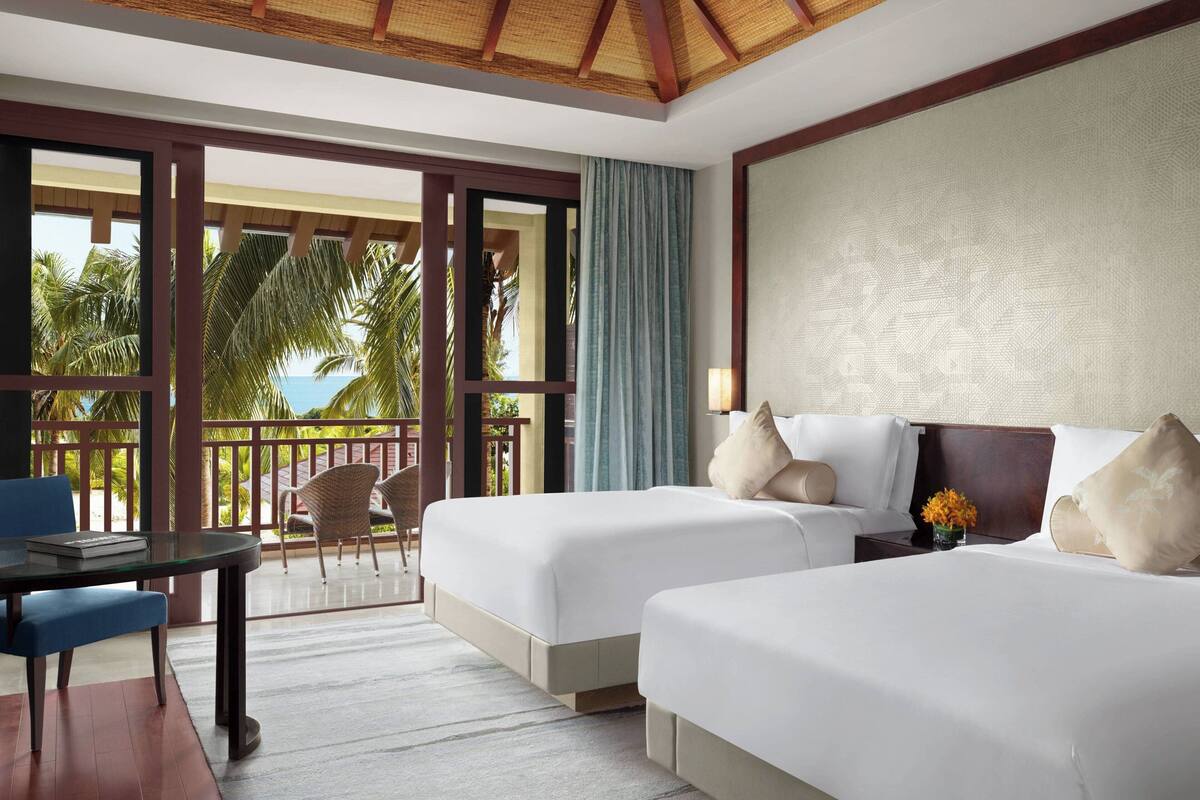 A hotel room with two beds, a balcony with a view of palm trees and the sea, a dining table with chairs, and a small table with a vase of flowers.