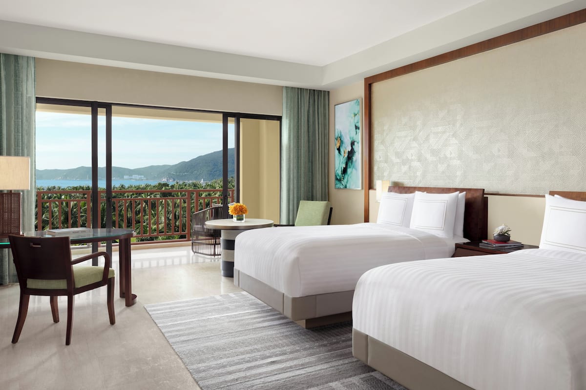 A hotel room with two beds, a balcony with a view of the sea, a desk, and a chair.