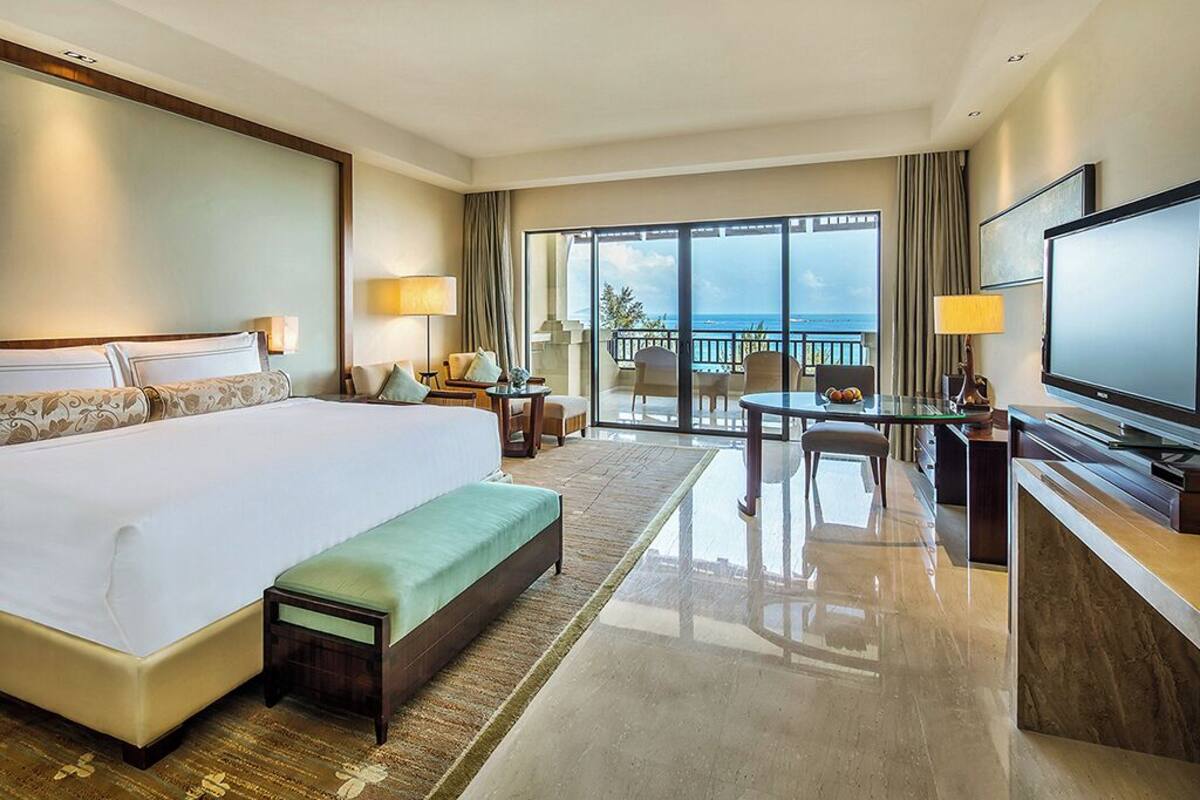 A hotel room with a large bed, a desk with a television, a dining area with chairs, and a balcony with a view.