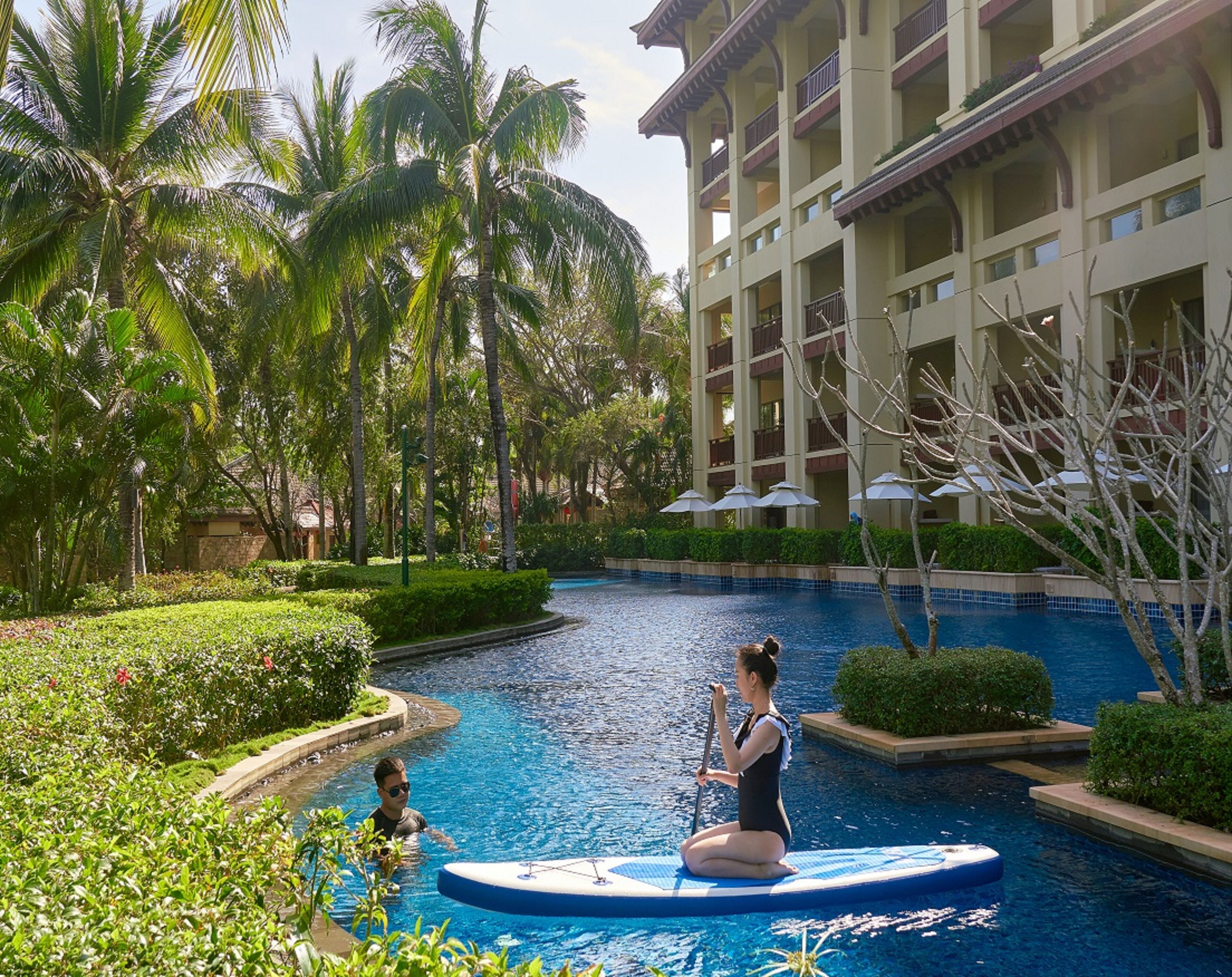 A swimming pool area with a person paddleboarding, surrounded by lush greenery and a multi-story building with balconies.