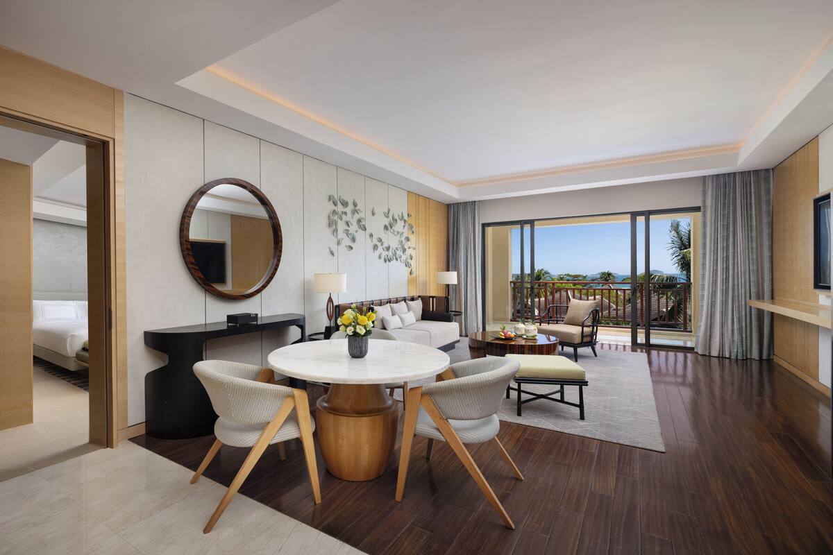 A modern hotel room with a dining area, a sofa set, a round table with chairs, a large mirror, and a view of the outdoors.