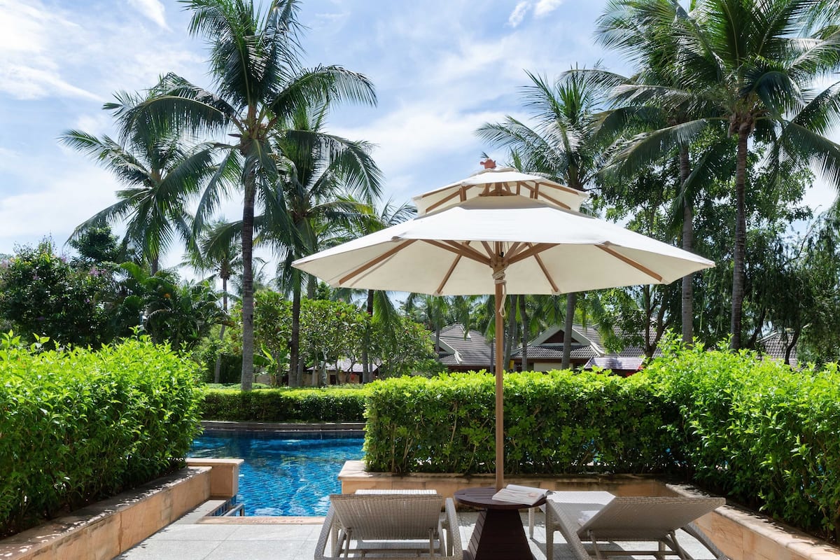 A poolside area with a table and chairs under a large umbrella, surrounded by lush greenery and palm trees.