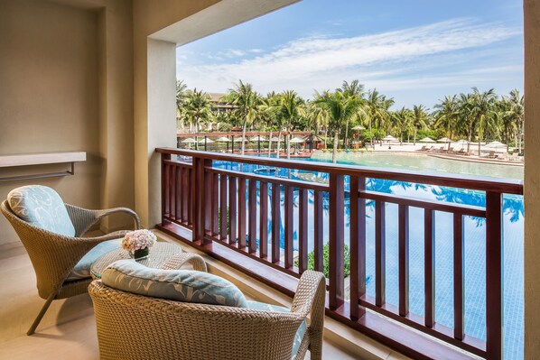 A balcony with wicker chairs and a view of a pool area with palm trees and lounge chairs.