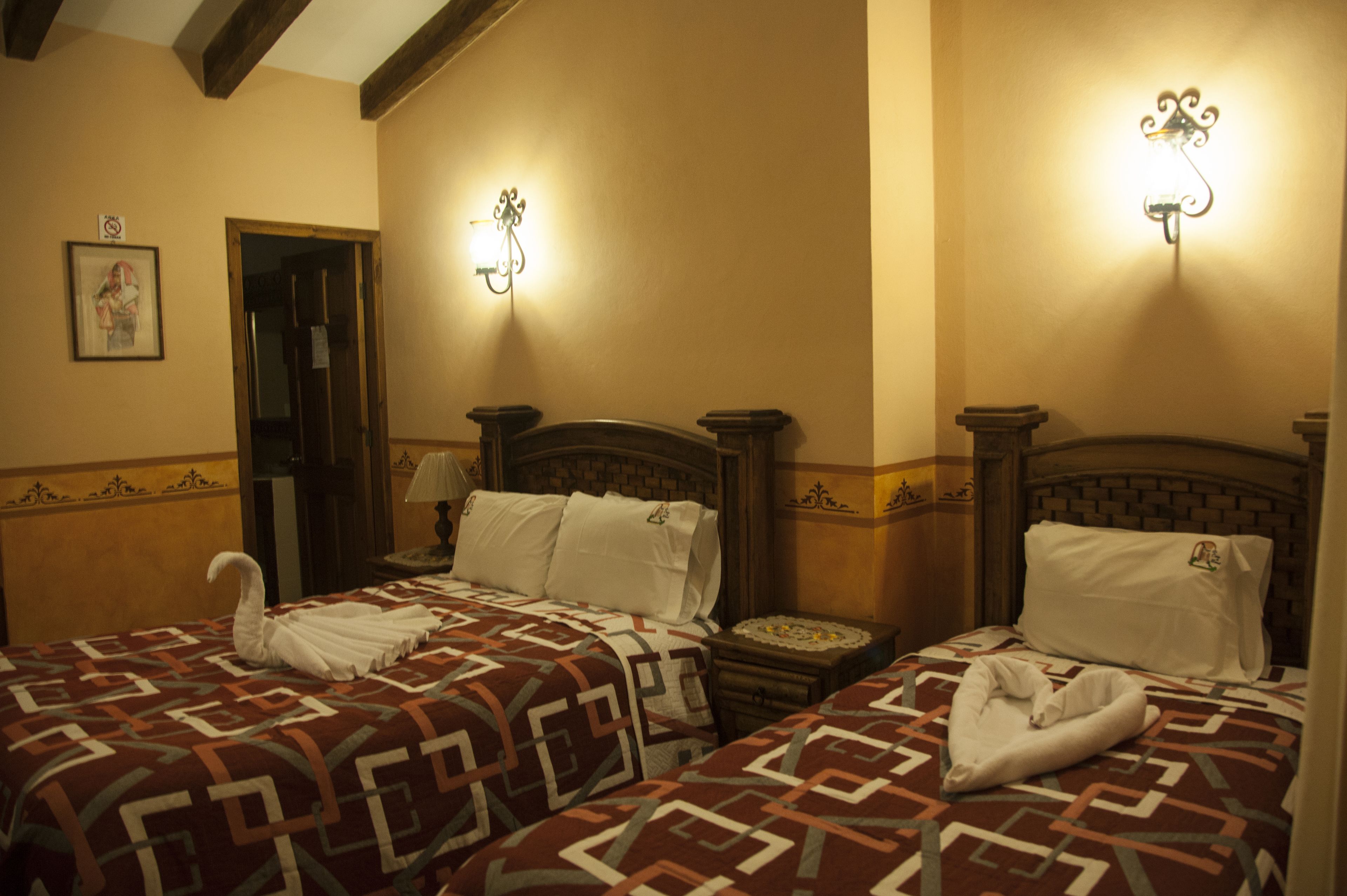 A hotel room with two beds, each with a patterned bedspread and white pillows. There are wall sconces providing light, and a framed picture on the wall.