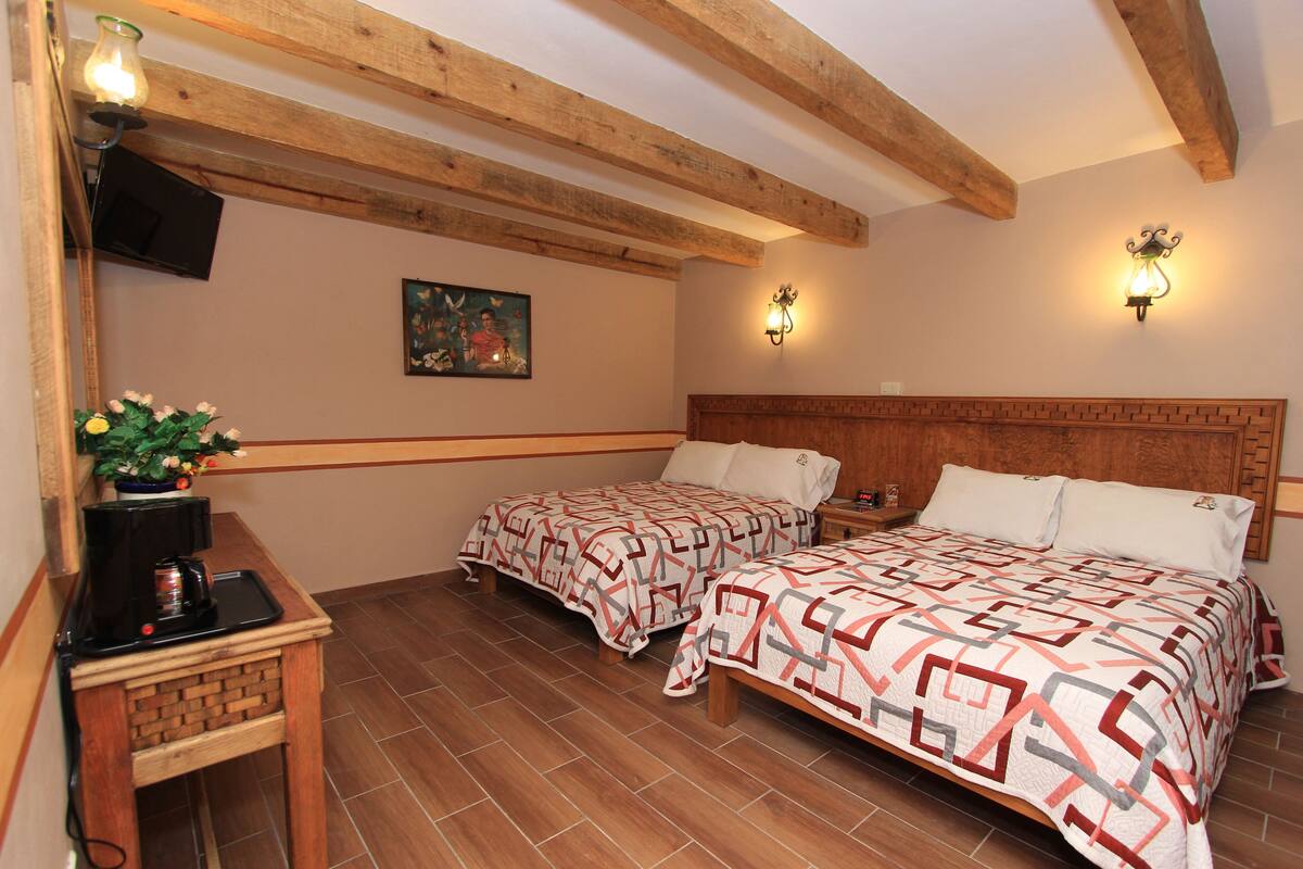 A room with two beds, wooden beams, a painting, wall sconces, a small table with a coffee maker, and a potted plant.