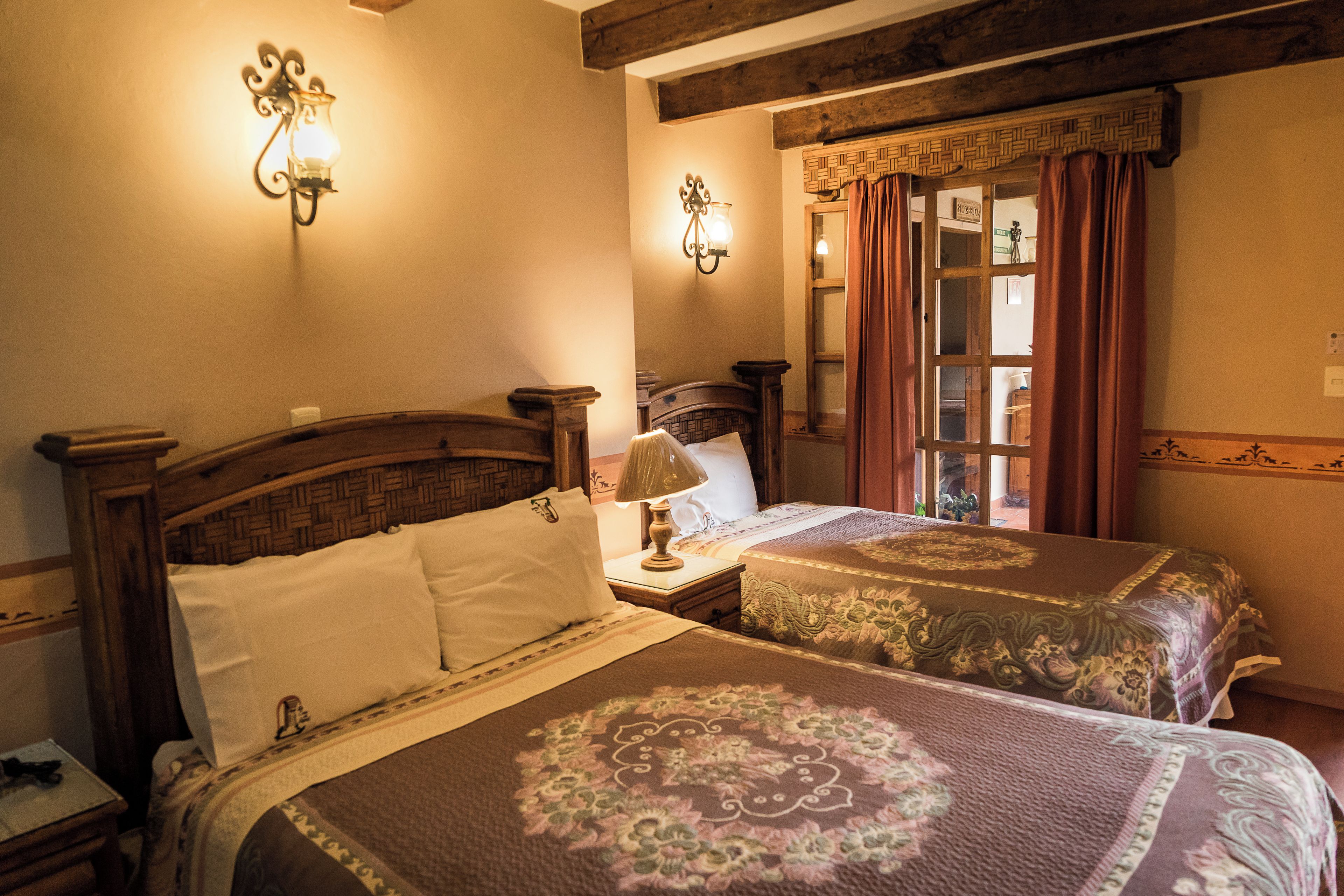 A hotel room with two beds, a nightstand, a lamp, and a window with curtains.