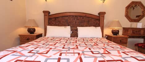 Una cama bien hecha con un edredón estampado, dos lámparas fijadas a la pared, un cabecero de madera y un espejo en la pared.