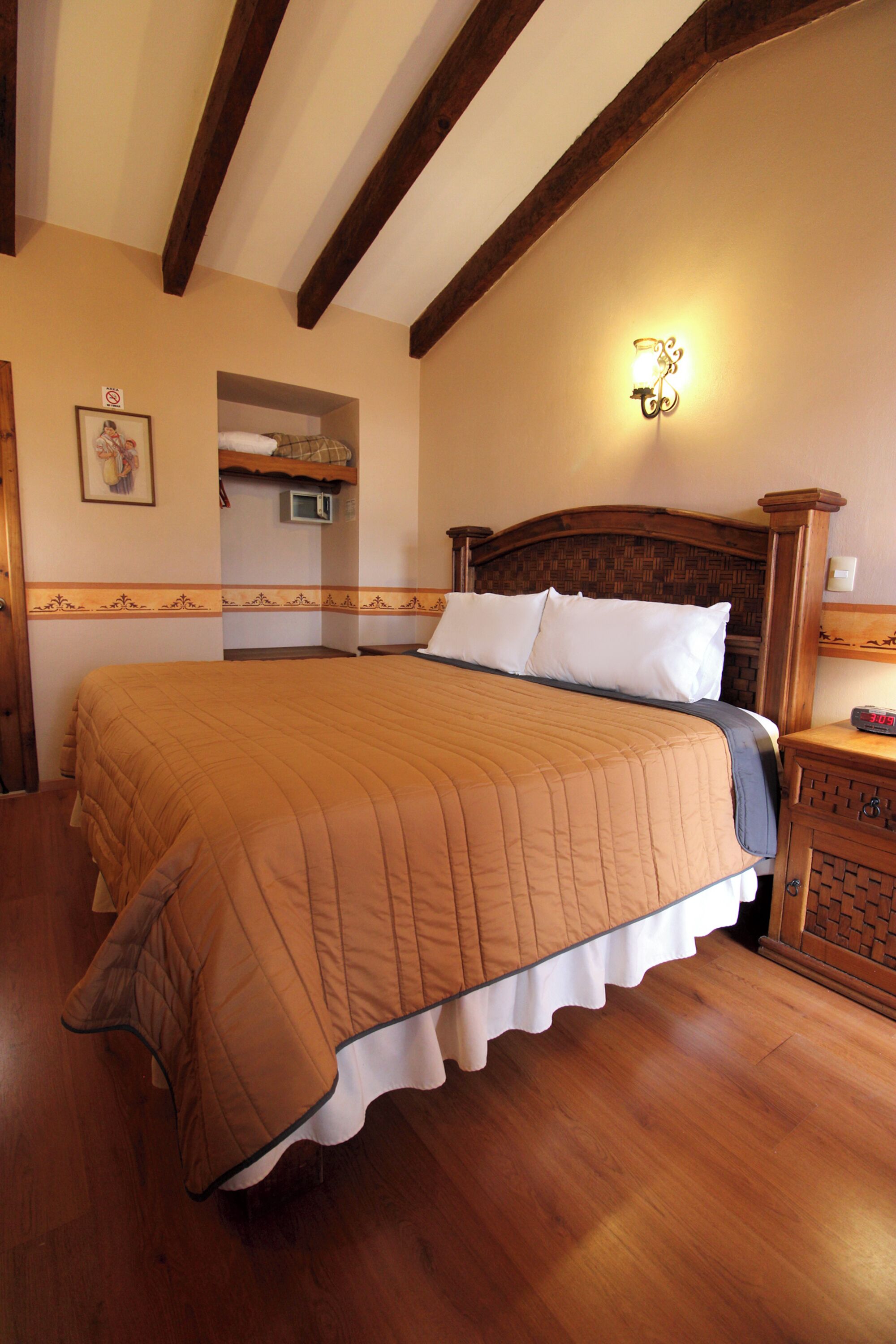 Un dormitorio con una cama grande, vigas de madera en el techo, una mesita de noche con lámpara, y un aplique de pared.