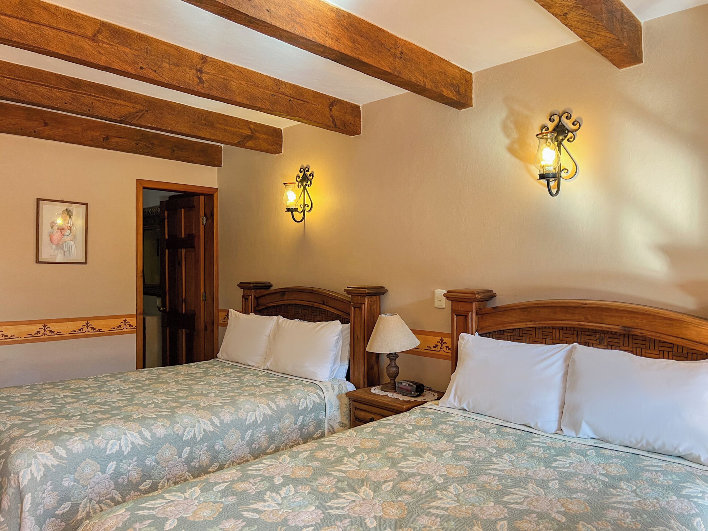 A hotel room with two single beds, wooden headboards, a nightstand with a lamp, and wall-mounted sconces.
