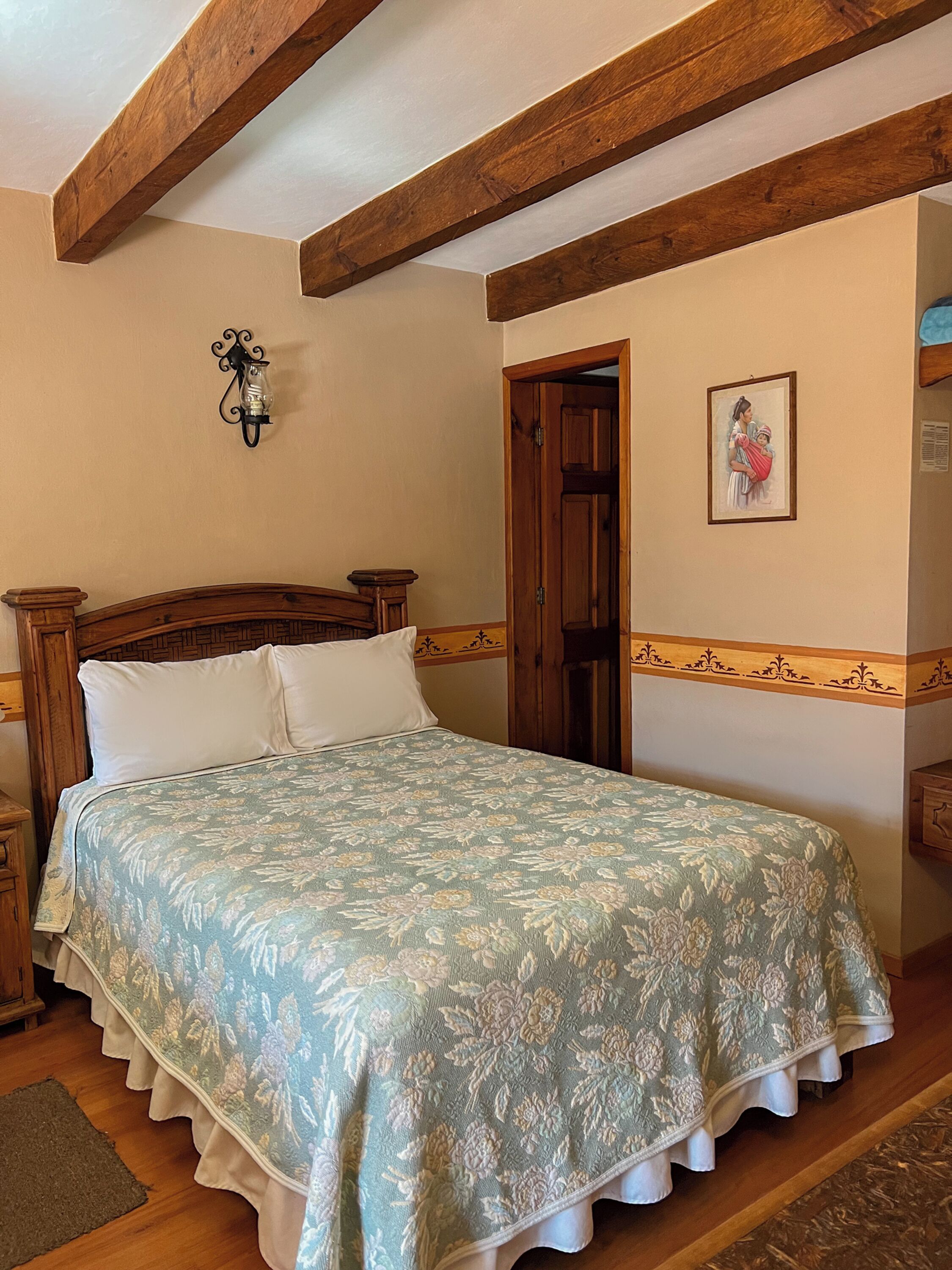 A bedroom with a wooden headboard, a floral bedspread, a nightstand, a wall-mounted lamp, and a door with a decorative panel.