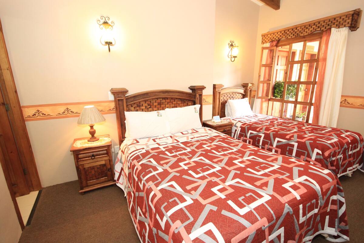 A hotel room with two beds, a nightstand with a lamp, and a window with curtains.