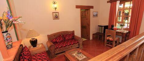 Una sala de estar acogedora con un sofá y una mesa de centro de madera, además de una lámpara. Se observa una ventana con cortinas y una puerta que da a otra habitación.