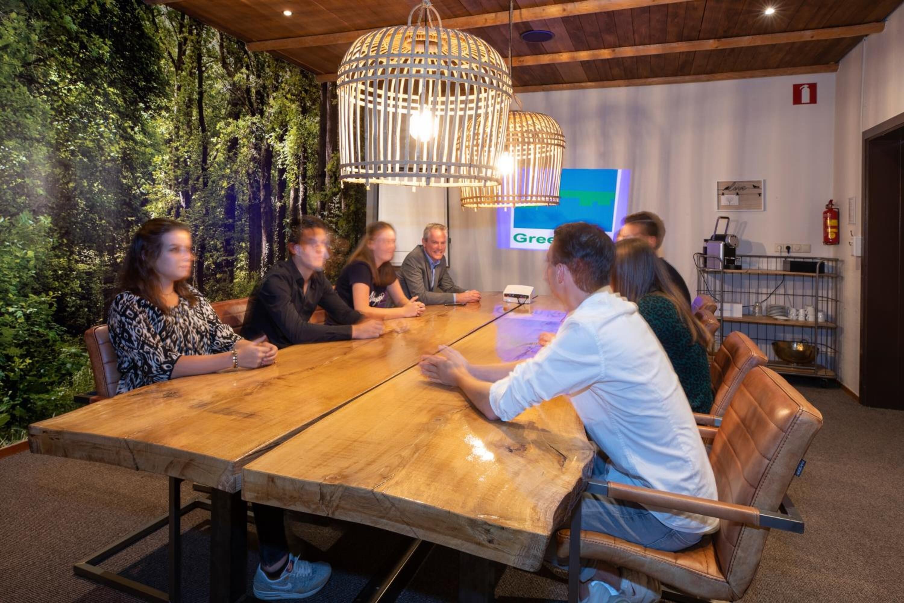 Eine Gruppe von Personen sitzt an einem Holztisch in einem Raum mit grüner Wand und einer Pendelleuchte.