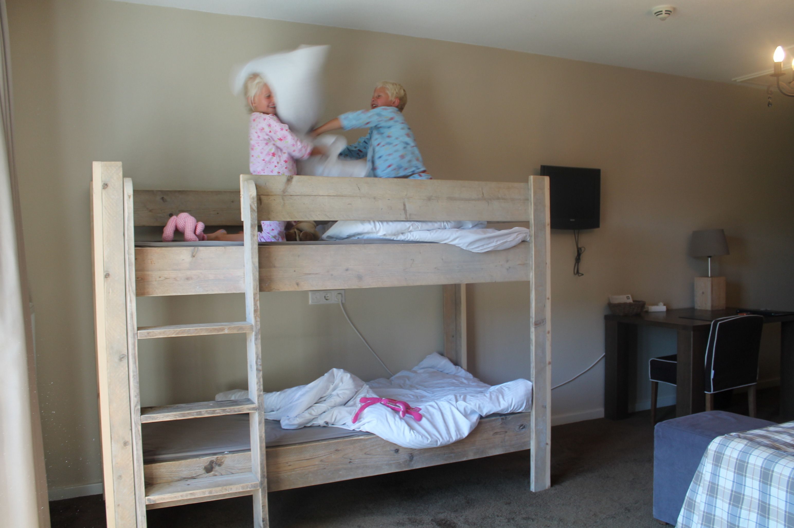 A bunk bed with two children playing on top. There is a desk with a chair and a lamp to the right.