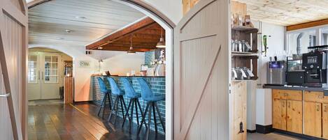 Un interior rústico con vigas de madera, una zona de bar con sillas azules y una cocina con una cafetera.