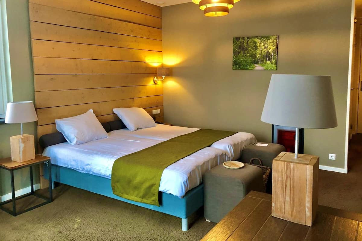 Un dormitorio con cabecera de madera, cama con ropa de cama blanca y un plaid verde, mesita de noche con lámpara, un pequeño otoman y un aplique de pared.