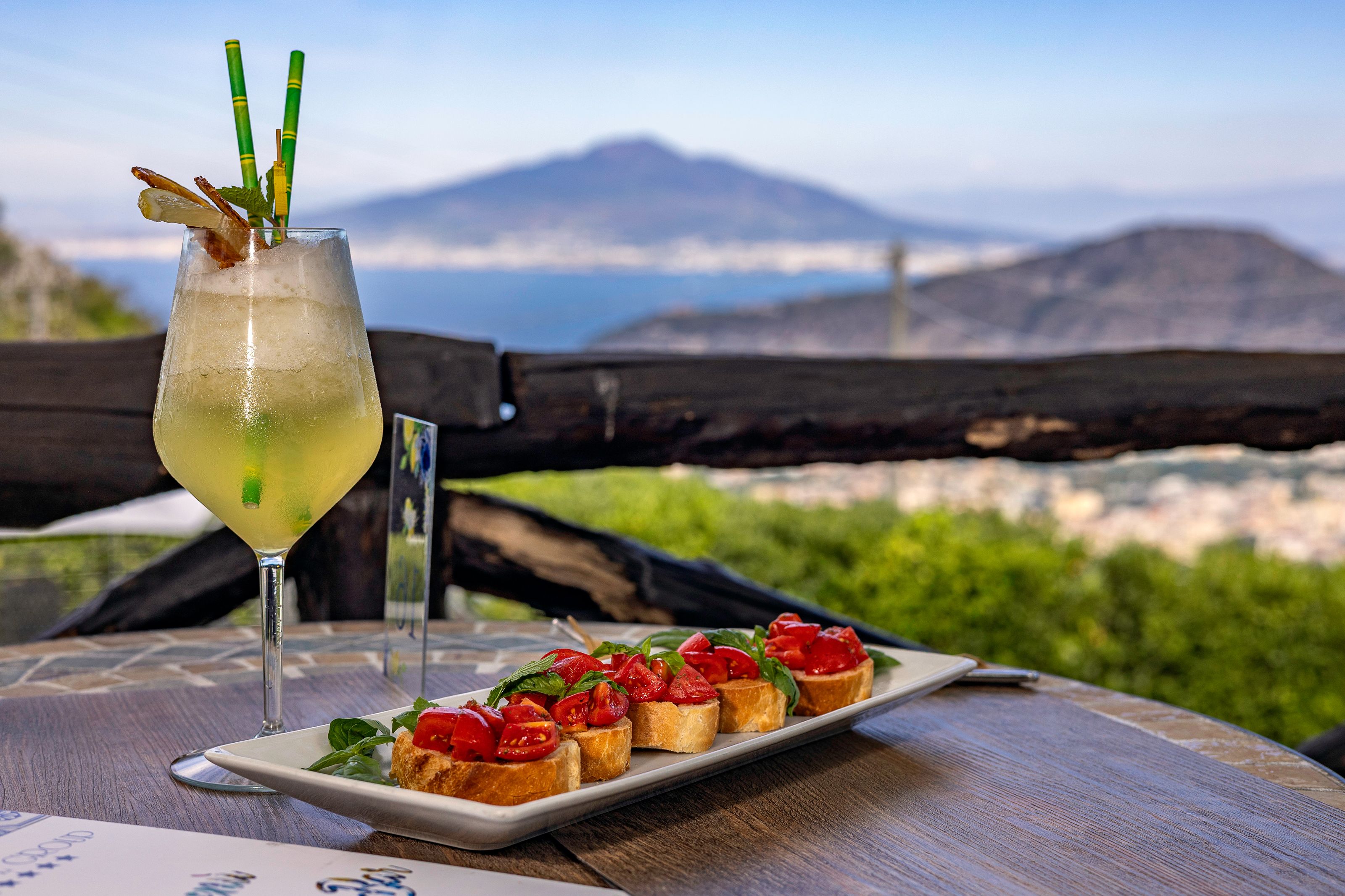 Une table sur laquelle se trouve une assiette de bruschettas et un verre d’une boisson jaune, agrémentée d’une paille et d’une garniture, le tout mis en valeur par un paysage montagneux en arrière-plan.