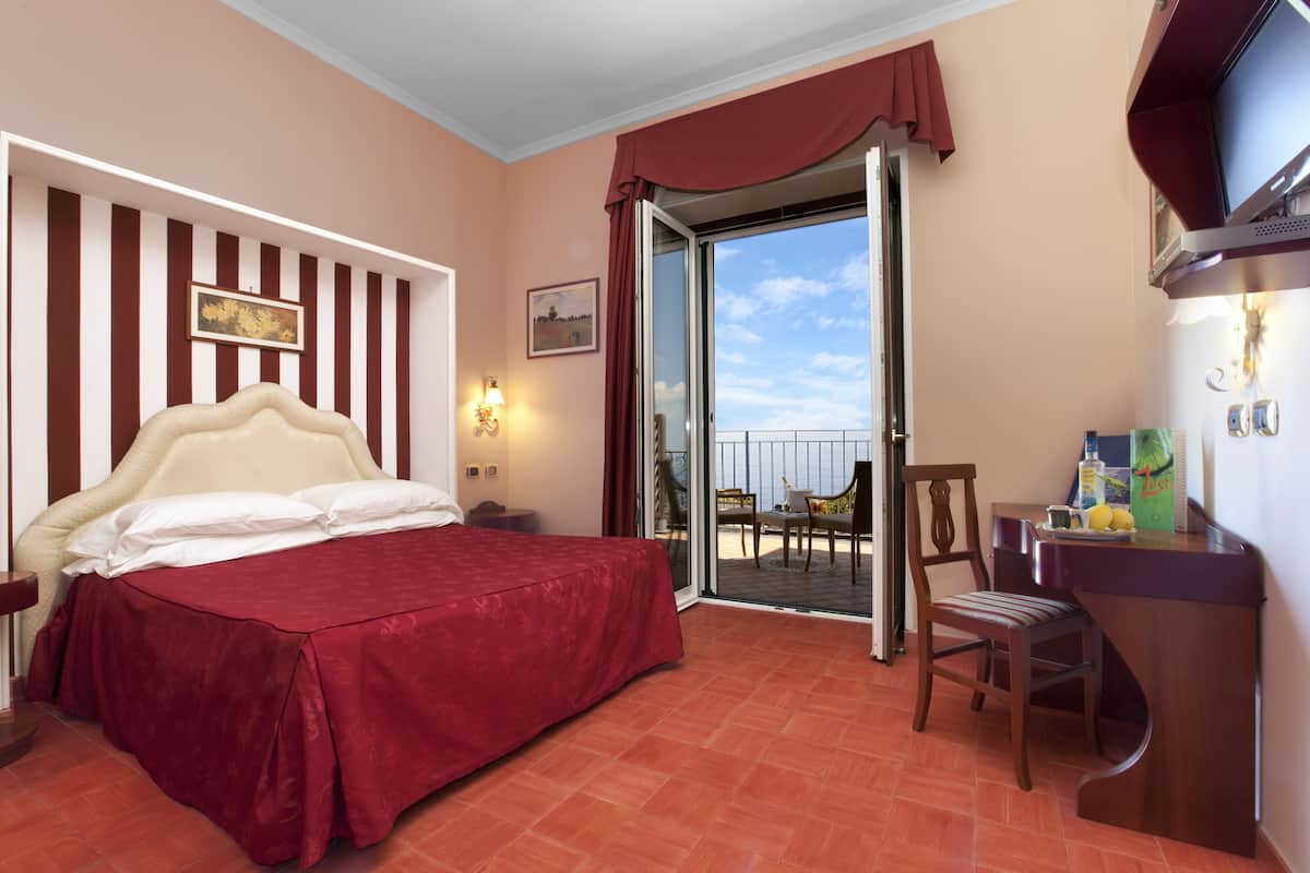 A hotel room with a bed, a desk, a chair, a TV, and a balcony with a table and chairs.