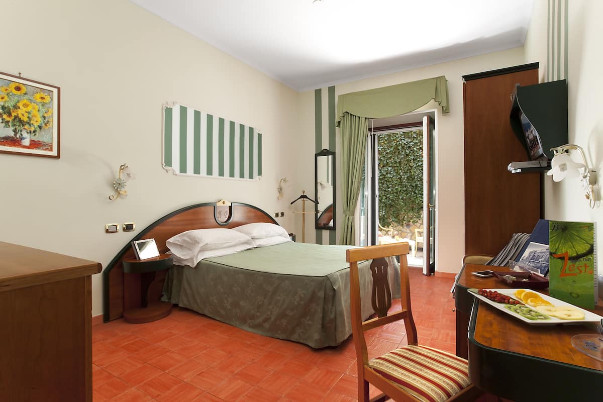 A hotel room with a bed, a desk, a chair, and a dining table with fruit.