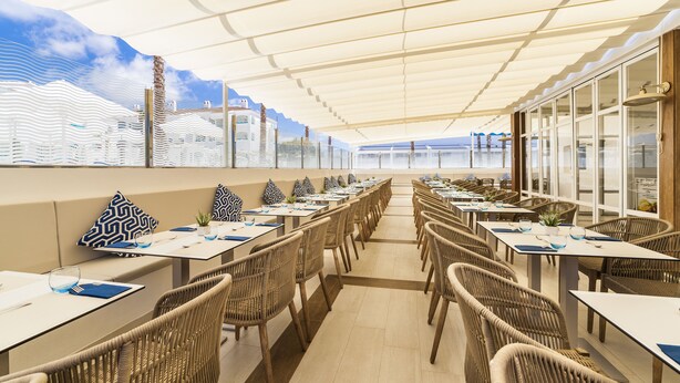 Un restaurante con sillas y mesas de mimbre, techo blanco y vista al mar.
