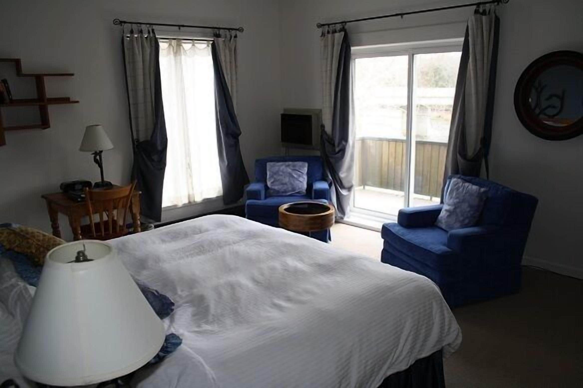 A bedroom with a bed, two blue armchairs, a small table with a lamp, a television, and a window with curtains.