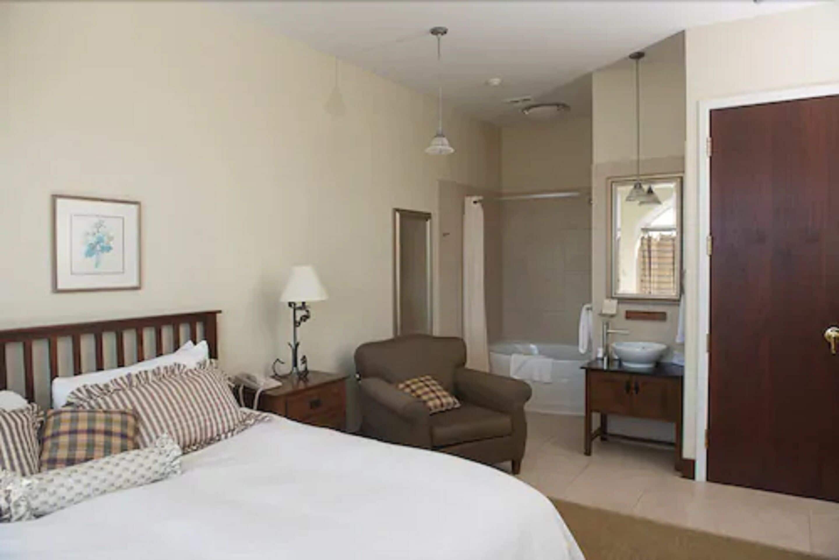 A hotel room with a bed, a chair, a nightstand, a lamp, a bathroom with a bathtub, and a sink.