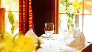 Une table à manger dressée avec une nappe blanche, des assiettes et des verres à vin, agrémentée d’un vase de fleurs jaunes et offrant une vue sur la fenêtre.