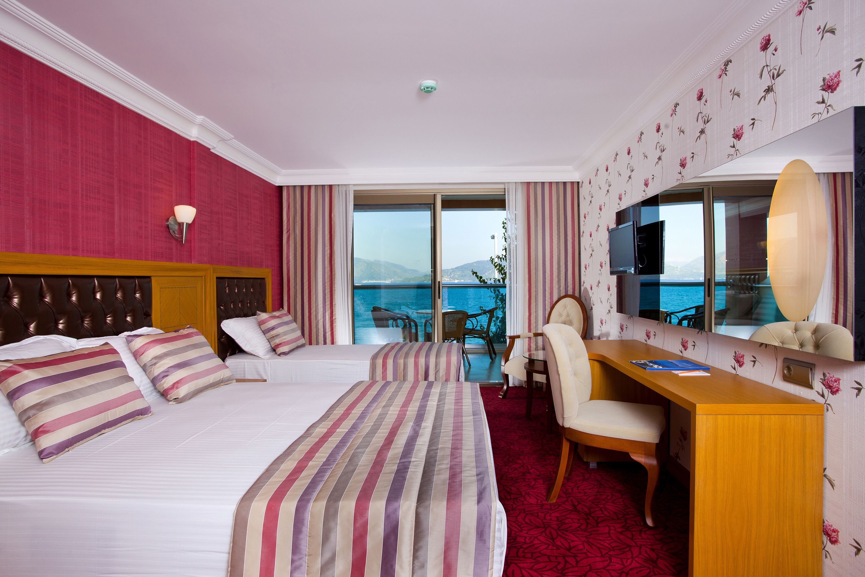A hotel room with two beds, a desk, a chair, a TV, and a large window with a view of the sea.