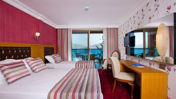 A hotel room with two beds, a desk, a chair, a TV, and a large window with a view of the sea.