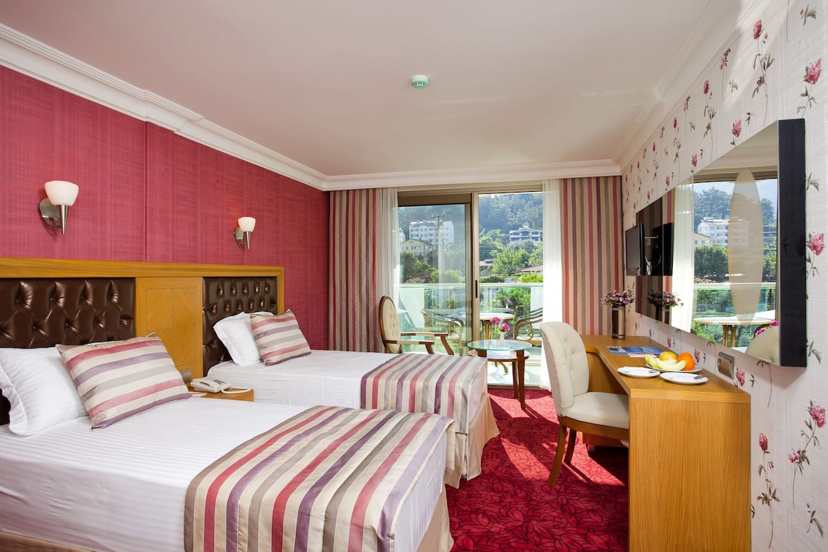 A hotel room with two beds, a desk with a chair, a TV, and a balcony with a view of buildings and trees.