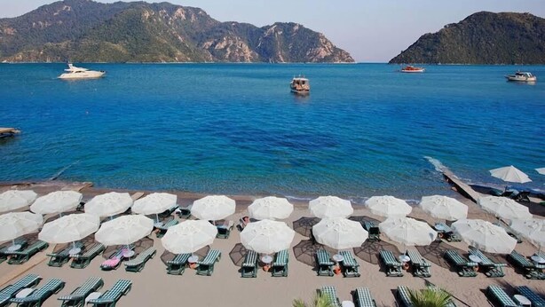 A beach with lounge chairs and umbrellas, boats on the water, and mountains in the background. - Orsmaris Boutique Hotel - Boutique Class (Marmaris)
