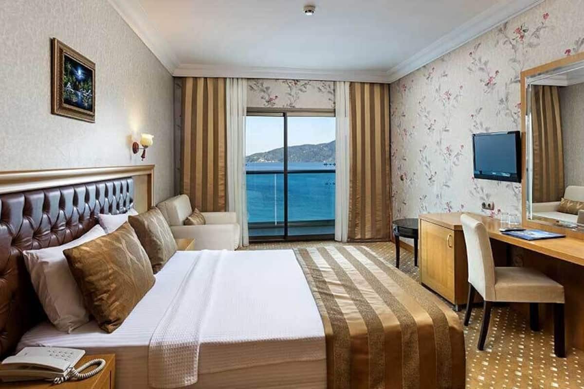 A hotel room with a bed, a desk, a chair, a television, and a view of the sea.