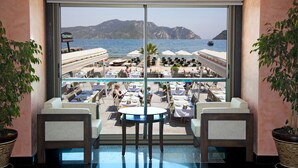 A view from a room with large windows overlooking a beachside restaurant with outdoor seating and a view of the sea and mountains. - Orsmaris Boutique Hotel - Boutique Class (Marmaris)