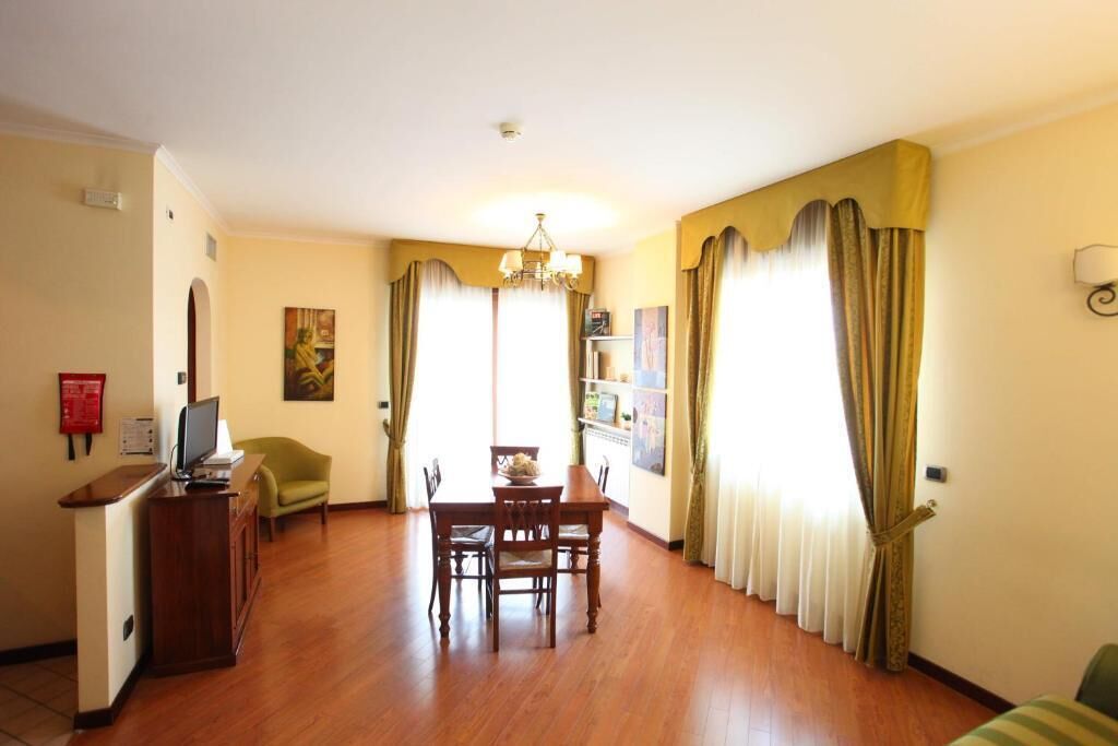 A hotel room with a wooden dining table, chairs, a small TV, and a desk. There are curtains on the windows, a chandelier, and a fire extinguisher on the wall.