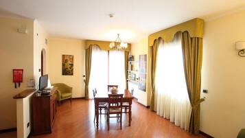 A hotel room with a wooden dining table, chairs, a small TV, and a desk. There are curtains on the windows, a chandelier, and a fire extinguisher on the wall.