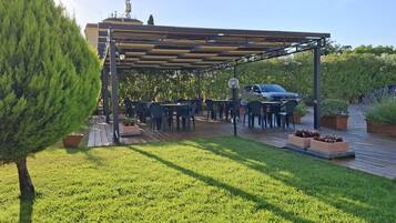Eine Terrasse im Freien mit Tischen und Stühlen, umgeben von Pflanzen und einem Gebäude im Hintergrund.